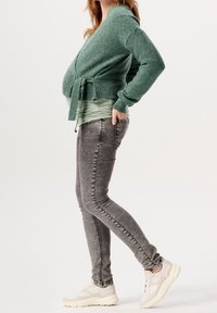 Grön stickad vikcardigan med knytning i midjan, lager på en ljusgrön topp, kombinerad med grå skinny jeans och vita sneakers.