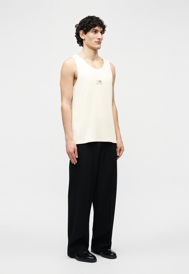 Uomo con capelli ricci che indossa una canottiera crema senza maniche e pantaloni neri larghi, in piedi contro uno sfondo chiaro e uniforme.