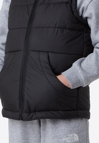 Svart vattert vest med glidelås foran og sidelommer, brukt over en grå genser. Materialet ser glatt ut med et quiltet design.