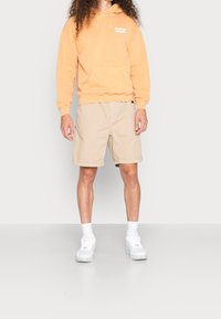 Orange hooded sweatshirt with a front pocket and white logo, paired with beige shorts and white sneakers. Model stands on a neutral background.