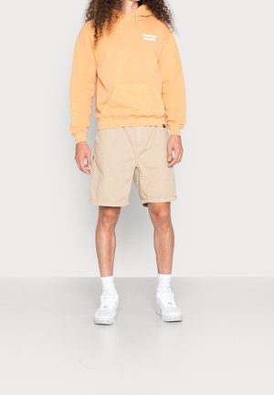 Felpa arancione con cappuccio, dotata di tasca frontale e logo bianco, abbinata a pantaloni corti beige e scarpe da ginnastica bianche. Il modello è in piedi su uno sfondo neutro.