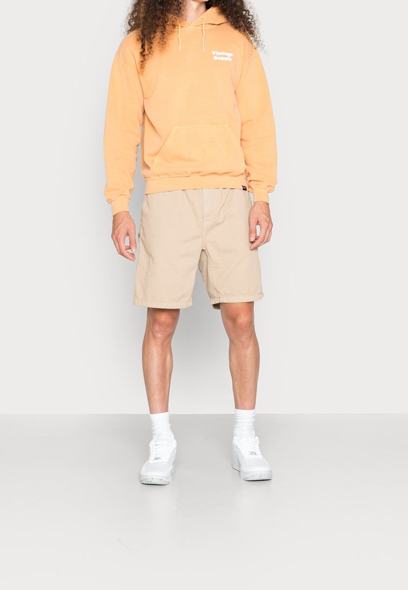 Orange hooded sweatshirt with a front pocket and white logo, paired with beige shorts and white sneakers. Model stands on a neutral background.