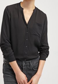 Person wearing a long-sleeve black button-up shirt with a chest pocket and faded black jeans, hands folded in front.