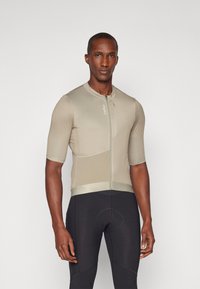 Kortermet sykkeltrøye i dempet beige med glidelås, teksturert stoff, og tettsittende design, paret med svarte sykkelshorts.