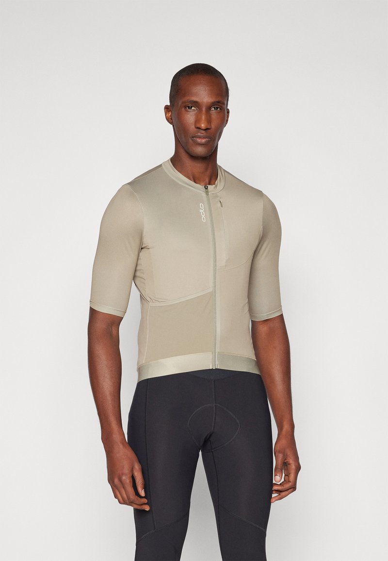 Kortermet sykkeltrøye i dempet beige med glidelås, teksturert stoff, og tettsittende design, paret med svarte sykkelshorts.