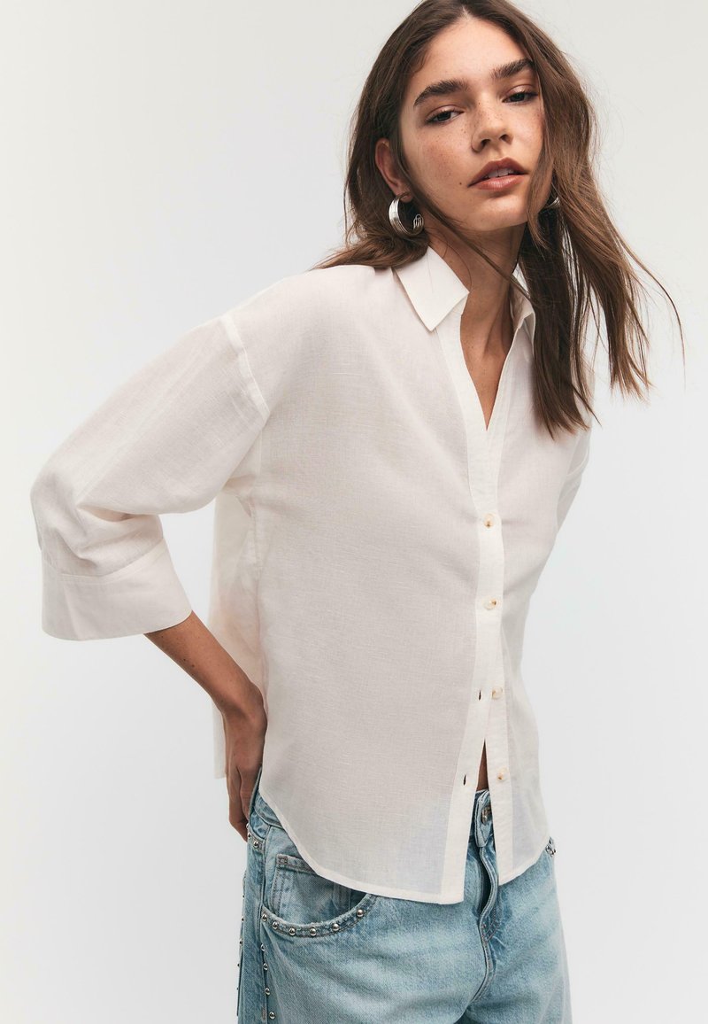 Jeune femme aux longs cheveux bruns portant une chemise blanche boutonnée et un jean bleu clair avec des détails à clous, posant devant un fond uni.