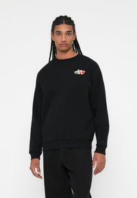 Sudadera negra de manga larga con puños acanalados. Presenta un pequeño logo bordado multicolor en el pecho. Diseño clásico de cuello redondo.