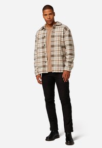 Beige und braun kariertes Flanelljackett mit Knopfverschluss, kombiniert mit einem beigen Hemd und schwarzen schmal geschnittenen Hosen. Schwarze Stiefel runden den Look ab.