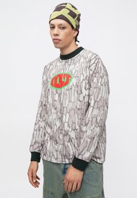 Tričko s dlhým rukávom v sivej farbe s textúrovaným, abstraktným vzorom; na hrudi má zelené a červené logo a čierne manžety.