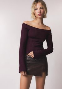 Bordeaux off-the-shoulder trui met geribbelde textuur en lange mouwen, gecombineerd met een bruin leren minirokje met studded accenten.