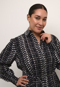 Abito nero con bottoni e un design a pattern bianco e grigio. Presenta un colletto, maniche lunghe e una vita legata, realizzato in un tessuto morbido.
