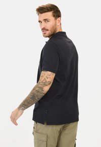 Sort polo shirt med klassisk krave, korte ærmer, sideslids og regular fit. Båret med olivengrønne cargobukser, der viser synlige tatoveringer.