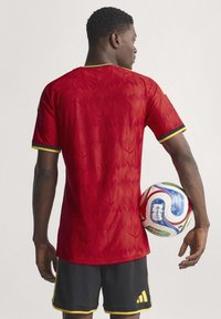 Crvena kratka nogometna majica s teksturiranim uzorkom, žutim obrubom i crnim akcentima. Drži šareni nogometni lopti.