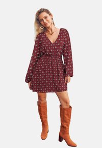 Bordeauxrotes Blumenkleid mit langen Ärmeln, V-Ausschnitt und elastischem Bund, kombiniert mit kniehohen braunen Wildlederstiefeln mit Blockabsatz.