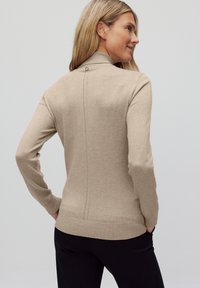 Beige stickad tröja med hög krage, långa ärmar och en subtil sömdetalj på baksidan. Ribbad nederkant och manschetter. Bärs över svarta byxor.