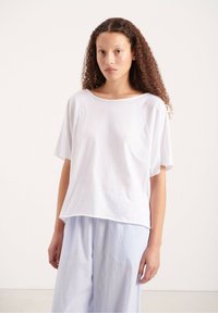 Balta, laisvai krentanti medvilninė marškinėliai su plačiu iškirpimu ir trumpomis raglan rankovėmis, derinami su šviesiai mėlynomis dryžuotomis kelnėmis.