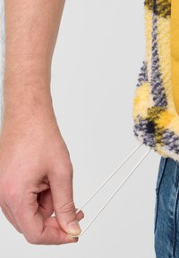 Una mano tiene due cordoni elastici bianchi attaccati a un capo di abbigliamento plaid giallo e grigio, morbido e con una superficie testurizzata.