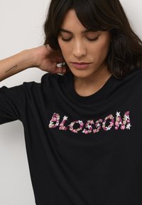 Čierne bavlnené tričko s kvetinovou výšivkou so slovom "blossom" v ružovej a bielej farbe, s okrúhlym výstrihom a voľným strihom.