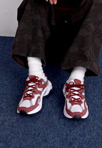 Scarpe da ginnastica rosse e grigie con upper in rete, suole in gomma e lacci. Indossate con calze bianche a metà polpaccio e pantaloni neri a fantasia. Sfondo di moquette blu.