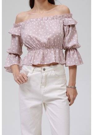 Vrouw draagt een roze off-shoulder polkadot top met ruches aan de mouwen en witte broek met hoge taille, poseert met één hand in haar zak.