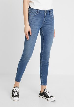 Personne portant un jean skinny bleu et des baskets basses noires, debout devant un fond blanc uni.