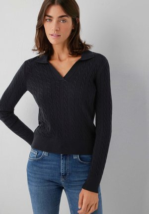 Donna che indossa un maglione nero a trecce a maniche lunghe con scollo a V e jeans blu, in piedi contro una parete chiara e semplice.