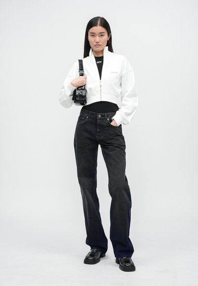 Giacca bianca cropped con zip sopra una maglietta nera, abbinata a jeans neri a gamba larga e scarpe nere; viene tenuta una piccola borsa nera strutturata.
