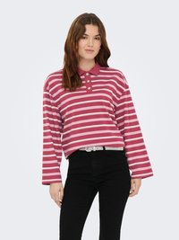 Polo à manches longues avec col, rayé rose et blanc, présentant une coupe décontractée et une patte de boutonnage à trois boutons, associé à un jean noir et une ceinture ornée de bijoux.