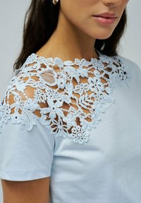 Camiseta azul clara con intrincados detalles de encaje azul claro en los hombros, patrones florales y una suave textura de algodón.