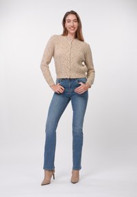 Cárdigan beige de punto con botones, combinado con jeans ajustados azules y tacones altos color nude. Textura suave y diseño entallado.