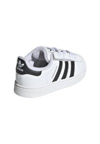 Baltā sporta apavi ar melnu trīs svītru dizainu, izgatavoti no ādas, ar teksturētu gumijas zoli un mīkstiem ieliktnis komfortam.