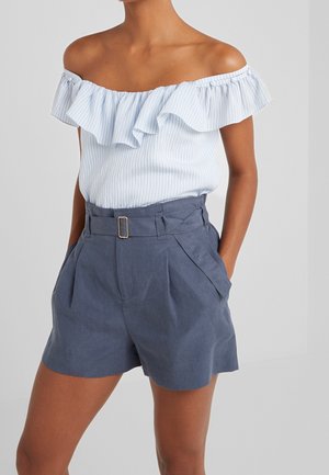 Vrouw draagt een lichtblauwe off-shoulder top met ruches, ingestopt in een met riem versierde, hooggetailleerde blauwe short met plooien en zijzakken.