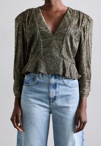 Metallisch olijfgroene top met een diepe V-hals, geplooide schoudernaden, peplum taille en textiel met structuur, gecombineerd met lichtblauwe high-waisted jeans.