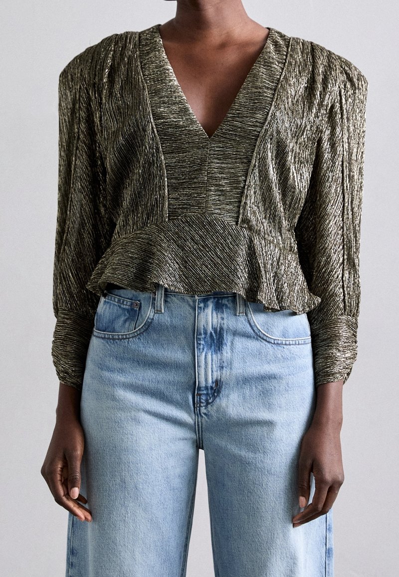 Metallisch olijfgroene top met een diepe V-hals, geplooide schoudernaden, peplum taille en textiel met structuur, gecombineerd met lichtblauwe high-waisted jeans.