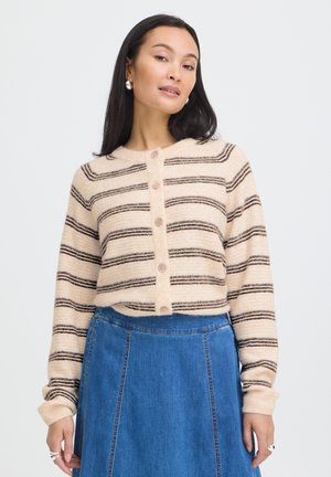 Kvinde står iført en beige og brunstribet cardigan med knapper, lange ærmer og en højtaljet blå denimnederdel.
