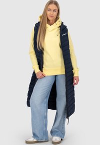 Vrouw in een lange marineblauwe gewatteerde vest over een gele hoodie, lichte blauwe wijde jeans en witte sneakers, staand tegen een effen achtergrond.
