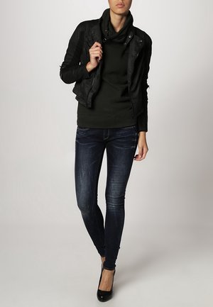 Chaqueta de cuero sintético negra con acentos acanalados, suéter verde oscuro, jeans oscuros ajustados y tacones altos negros. Conjunto simple y moderno.