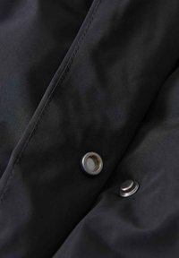 Dettaglio di una giacca in tessuto nero con texture liscia, caratterizzata da due bottoni a pressione metallici. Cuciture lineari visibili lungo i bordi.