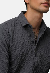 Camisa de botones gris con una tela texturada y estampada que presenta un diseño en forma de rejilla y un collar, complementada por botones oscuros.