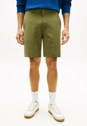 Mann trägt kniehohe olivgrüne Shorts, weiße Socken und weiße Turnschuhe, steht vor einem schlichten hellen Hintergrund.