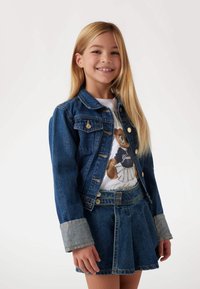 Jeansjacka, blå med knappar och bröstfickor, tillsammans med en jeanskjol med veckad design; modell med långt hår och T-shirt under.