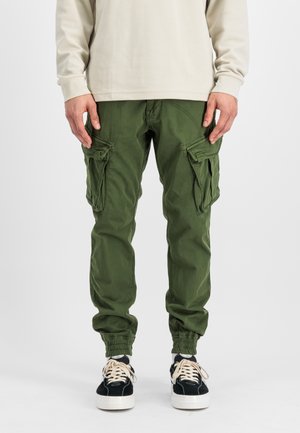 Man wearing green cargo pants with elastic cuffs and black sneakers with white soles and laces, standing against a plain background.