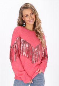 Różowy sweter z długimi, błyszczącymi frędzlami w kształcie litery V. Dzianinowa tekstura, luźny krój i ściągacze na mankietach. Nosi się go na niebieskie dżinsy.