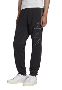 Černé tepláky s elastickými manžetami, dvěma bočními kapsami a tónovým zigzagovým vzorem na levé noze. Vyrobeny z měkkého, flexibilního materiálu.