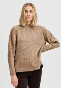 Brauner Strickpullover mit hohem Kragen, lockerer Passform und langen Ärmeln. Weiche Textur und leicht meliertes Material, kombiniert mit dunklen Hosen.