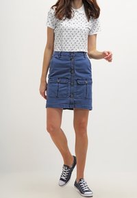 Mujer vestida con una camisa polo blanca con un patrón, una falda azul de botones con bolsillos y zapatillas bajas negras, de pie contra un fondo blanco.