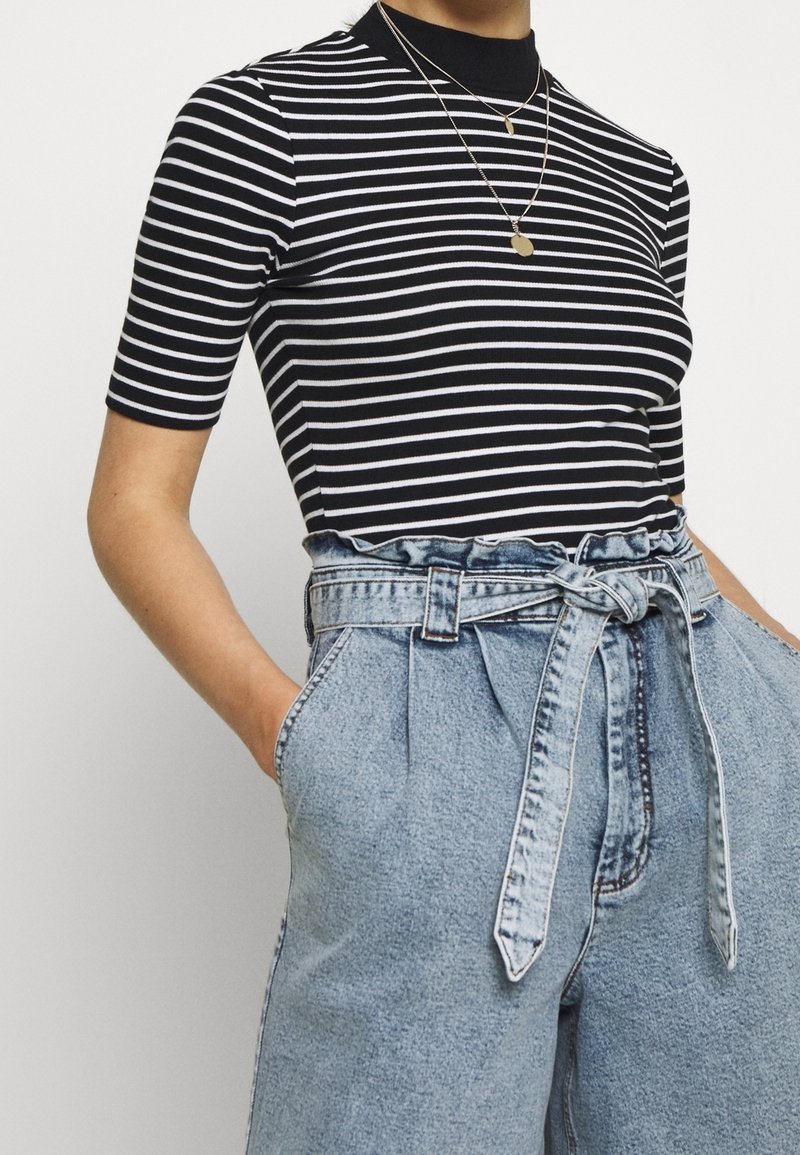 Personne portant un haut à manches courtes rayé noir et blanc et un pantalon en denim bleu clair taille haute avec ceinture nouée, main dans la poche.