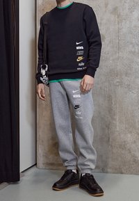 Persona con sudadera negra y pantalones deportivos grises con múltiples logotipos de Nike, zapatillas negras y llevando una pequeña bolsa peluda en blanco y negro.