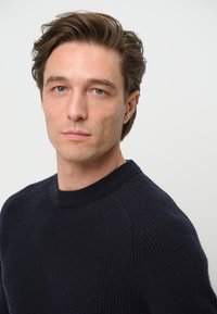 Marinblå ribbad tröja med rundad hals, texturerad stickning och figurnära passform, visad på en modell mot en enkel vit bakgrund.