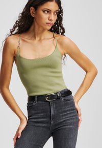 Top de tip fitted, de culoare verde măsliniu deschis, cu un finisaj texturat, bretele subțiri și accente din metal argintiu, asociat cu blugi gri închis și o curea neagră.